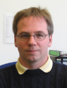 Sing mit Brille, dunklem Pullover mit hellem Kragen, neutraler Gesichtsausdruck.